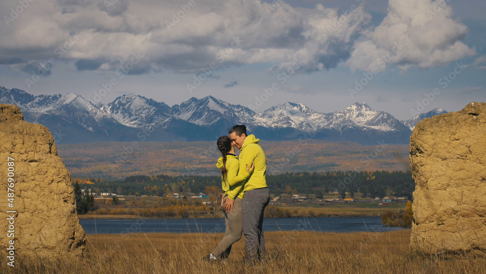Man and woman in yellow green sportswear. Lovely couple of travelers hug and kiss near old stone enjoying highland landscape. Two travelers are walking against the backdrop of snow-capped mountains.