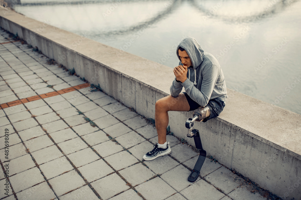 A handicapped sportsman with artificial leg with hooded sweatshirt sitting at the quay and warming up hands. A handicapped sportsman with artificial leg with hooded sweatshirt sitting at the quay and warming up hands.