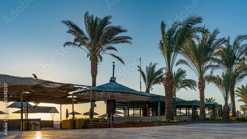 A paved pedestrian path runs along the beach. The rising sun shines through the lattice umbrellas and awnings. Palm trees against a blue sky. The Red Sea. Egypt