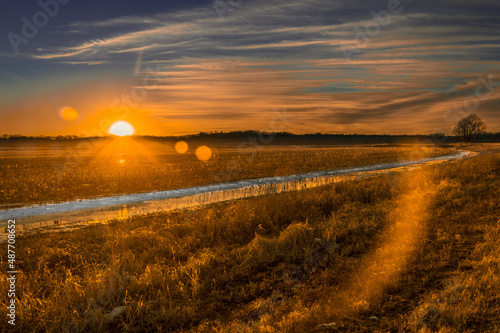 Beautiful sunset over dry agricultural field; narrow creek edged with snow crosses the field; winter in Midwest