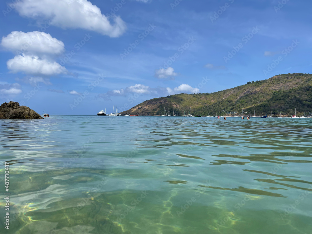 Clear turquoise sea water. Phuket, tropical paradise, Thailand. Headland, covered by dry and green grass, bushes and palm trees. Swimming people and sailboats at the distance. Anchored yachts, sea bay