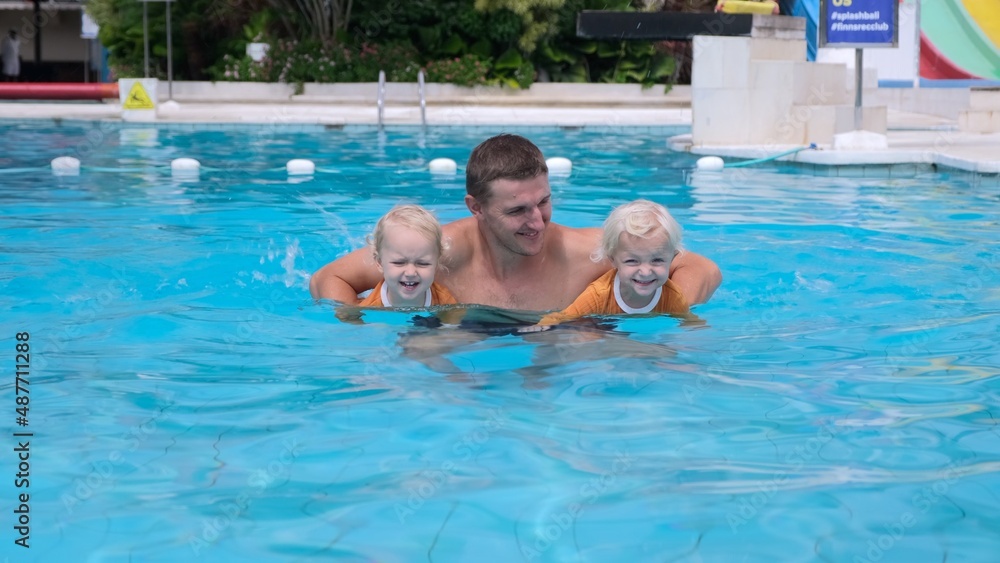Smiling dad with twin toddler daughters, teaching kids to swim in pool, happy family holiday vacation