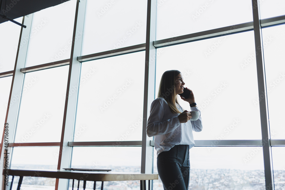 Young businesswoman wearing glasses and holding a phone. In an office or business center against the background of large windows of a skyscraper. Work and career, concept Young businesswoman wearing glasses and holding a phone. In an office or business center against the background of large windows of a skyscraper. Work and career, concept