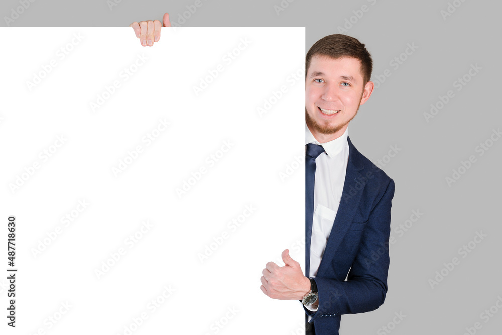 A businessman shows a thumbs up gesture holding onto a white blank board while standing on a gray background A businessman shows a thumbs up gesture holding onto a white blank board while standing on a gray background