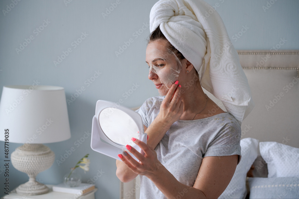 Nice woman with towel on her head applies moisturizing mask to her face and looks into a small mirror. After-shower care to preserve the beauty and health of the skin Nice woman with towel on her head applies moisturizing mask to her face and looks into a small mirror. After-shower care to preserve the beauty and health of the skin