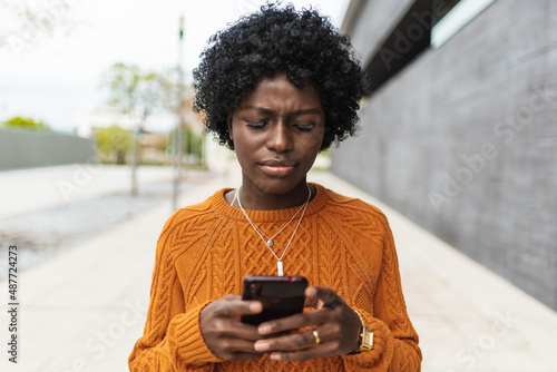 Young afro woman looks confused while watching something on her mobile phone outdoors in the street. Technology concept.