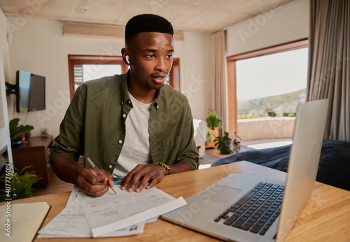 Young adult African American male wearing earphones making notes while on online meeting. Focussed, sitting at kitchen counter using laptop.