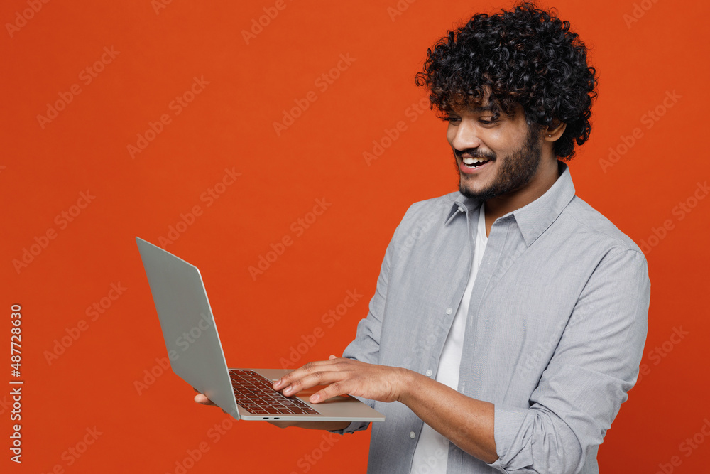 Smiling happy vivid young bearded Indian man 20s years old wears blue shirt hold use work on laptop pc computer typing browsing chatting send sms isolated on plain orange background studio portrait. Smiling happy vivid young bearded Indian man 20s years old wears blue shirt hold use work on laptop pc computer typing browsing chatting send sms isolated on plain orange background studio portrait.