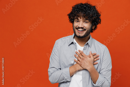 Happy good charming kind fascinating surprised young bearded Indian man 20s years old wears blue shirt ask who me oh it so sweet put hands on chest isolated on plain orange background studio portrait. © ViDi Studio