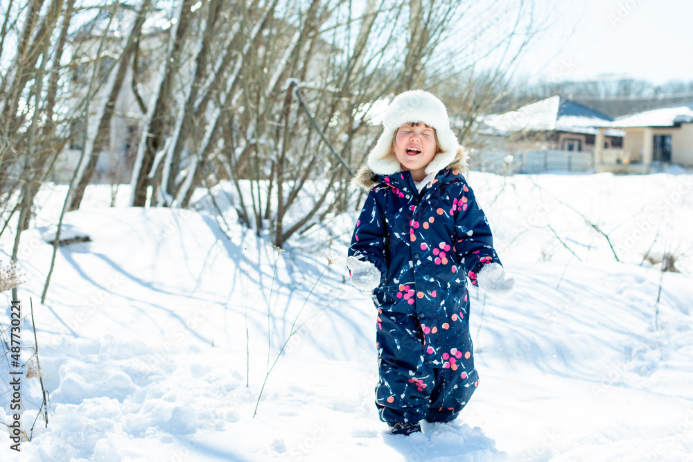 a funny little girl in overalls and a hat catches snow with her tongue in winter outside