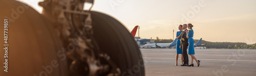 Full length shot of male pilot posing for photoshoot together with two air hostesses in blue uniform, standing in an airport terminal at sunset. Aircraft, aircrew concept photo