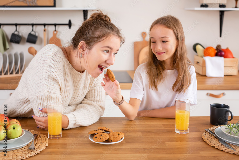 A young happy beautiful family has breakfast together at home. two cute sisters eat delicious cookies, healthy food in the morning, talk and smile while sitting in a modern kitchen A young happy beautiful family has breakfast together at home. two cute sisters eat delicious cookies, healthy food in the morning, talk and smile while sitting in a modern kitchen