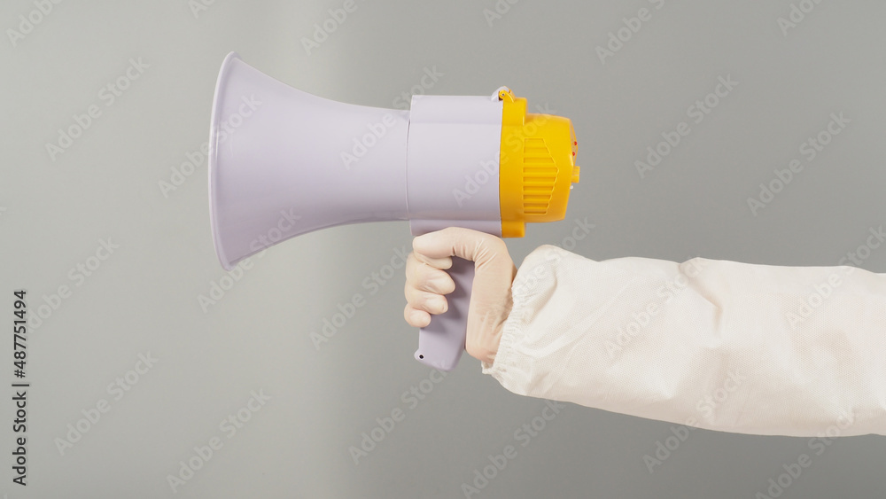 Hand is holding megaphone on grey background. Hand wears a PPE suit and a white medical glove. Hand is holding megaphone on grey background. Hand wears a PPE suit and a white medical glove.