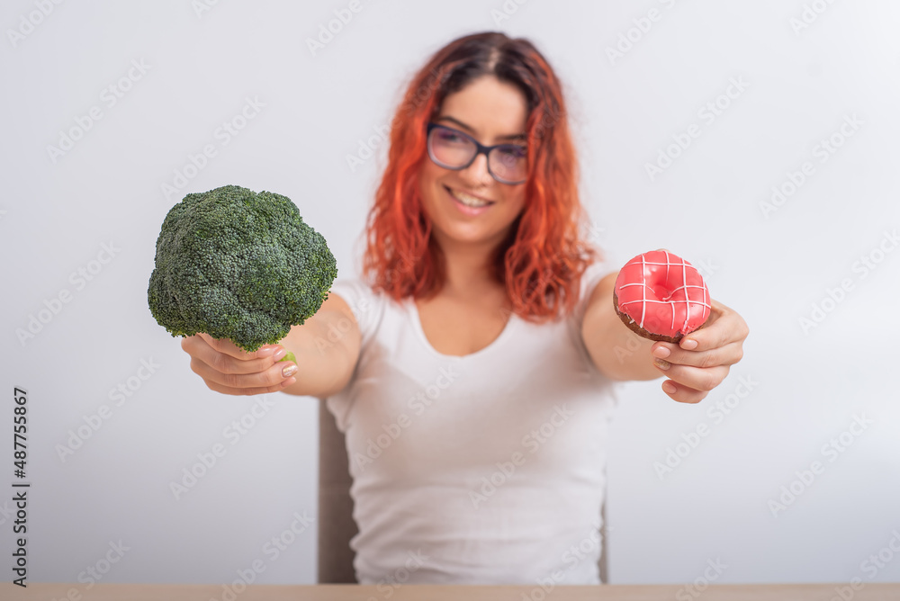 Caucasian woman chooses between vegetables and fast food. Redhead girl holding broccoli and donut on a white background. Caucasian woman chooses between vegetables and fast food. Redhead girl holding broccoli and donut on a white background.
