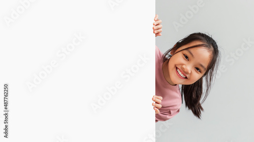 Cheerful Asian Schoolgirl Holding Empty Paper Poster, Gray Background