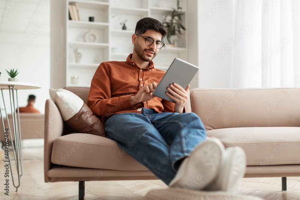 © Prostock-studio - Smiling young Arab man using digital tablet at home © Prostock-studio - Smiling young Arab man using digital tablet at home