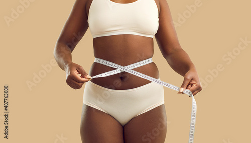 Close up crop of fat African American woman in underwear on yellow studio background measure waist with tape. Overweight black girl make measurement before diet. Weight loss and body shape. photo