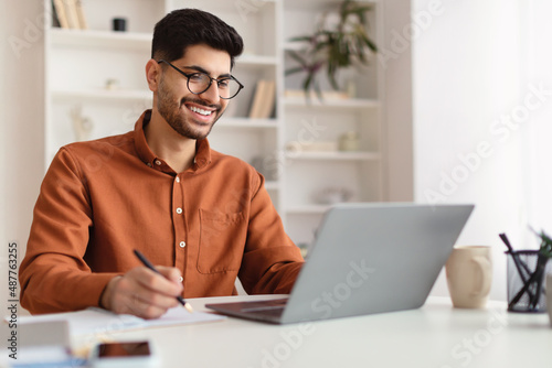 Smiling Arab man in glasses using laptop and writing photo