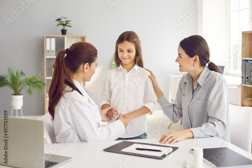 Little teen girl during medical examination receives support from her mother and friendly female pediatrician. Doctor holds child's hands and mother touches shoulder, assuring that everything is fine.