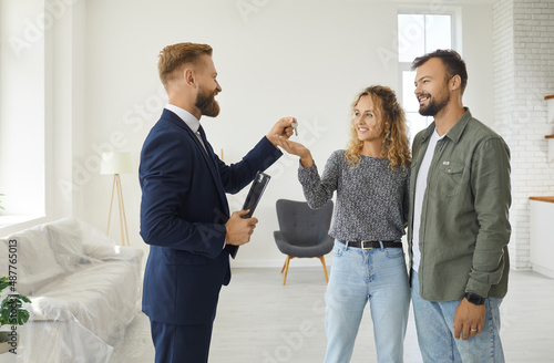 Real estate agent gives house keys to young married couple. Happy boyfriend and girlfriend or husband and wife standing in living room of new partially furnished apartment take keys that realtor gives photo