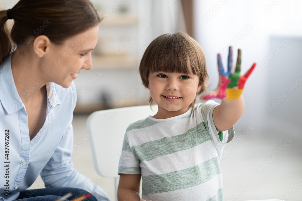 Happy little boy showing painted colorful hand and smiling to camera, sitting at school with tutor Happy little boy showing painted colorful hand and smiling to camera, sitting at school with tutor