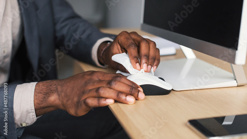 Unrecognizable black office worker wiping computer mouse with disinfectant at desk, closeup