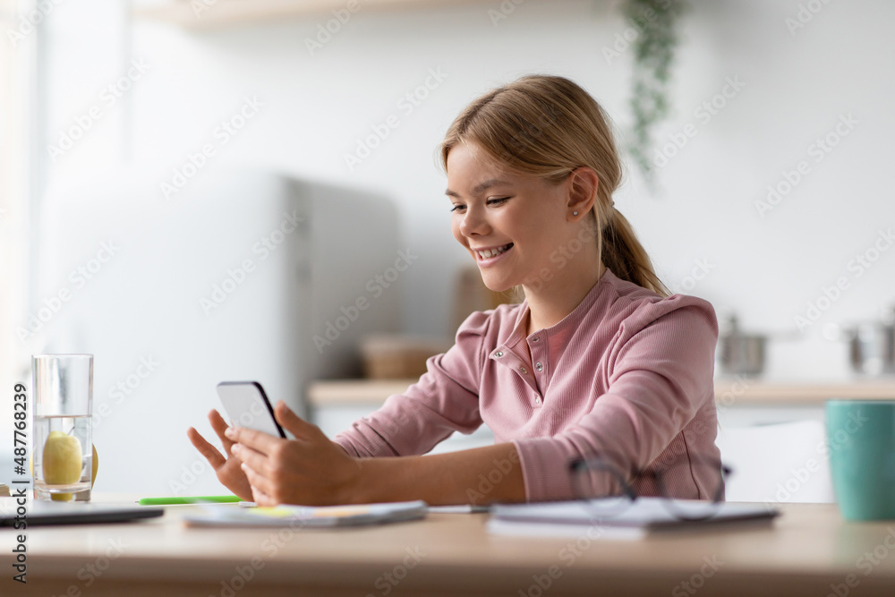 Glad smiling european blond youngsters girl reads message, chatting on smartphone in kitchen interior Glad smiling european blond youngsters girl reads message, chatting on smartphone in kitchen interior