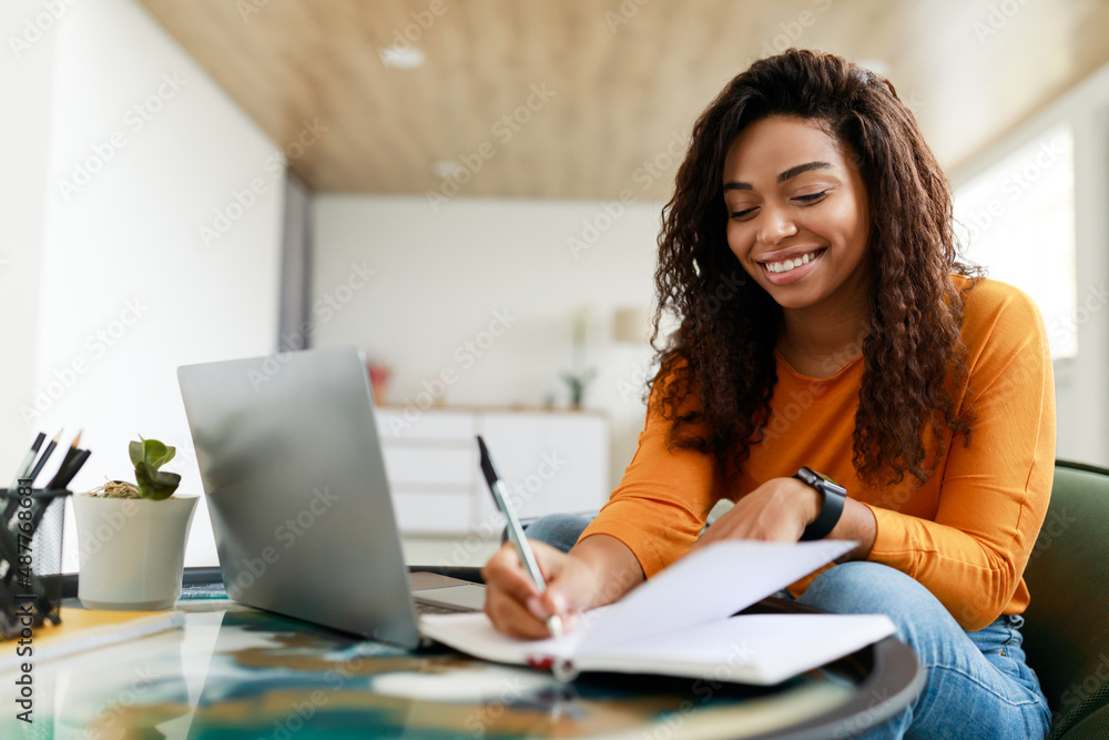 Black woman sitting at desk, using computer writing in notebook Black woman sitting at desk, using computer writing in notebook