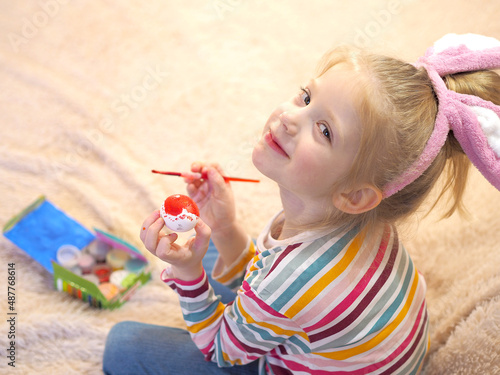 A happy little blonde girl with bunny ears laughs and paints an Easter egg with a paintbrush. Easter preparation. Making Easter decor.