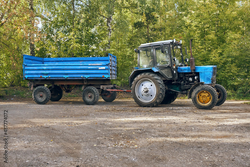 A blue agricultural tractor with a trailer on a dirt road, against a backdrop of bushes with green leaves. Special equipment for different types of work