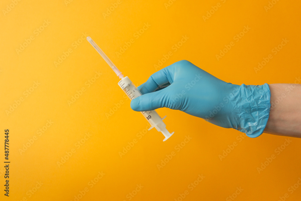 Man's hand in blue surgical glove holds a syringe with injection dose on the yellow background. Vaccination concept. Man's hand in blue surgical glove holds a syringe with injection dose on the yellow background. Vaccination concept.