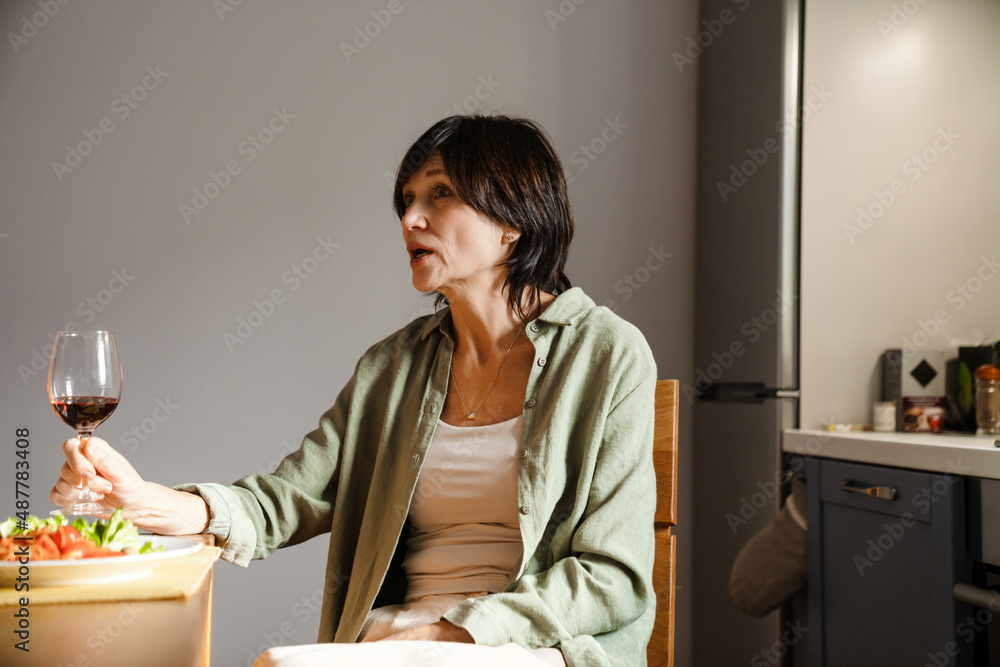 Mature brunette woman drinking wine while having dinner in kitchen Mature brunette woman drinking wine while having dinner in kitchen