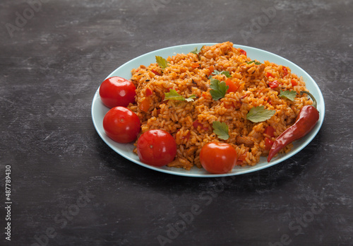 Jollof rice, tomatoes and hot peppers on a blue plate on a black background. National cuisine of Africa.
