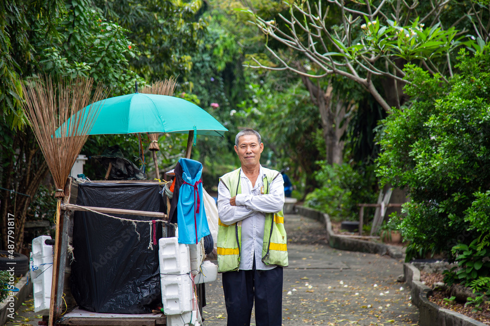 Portrait happy old Asian man street cleaner standing with his arms crossed next to an old gabage cart before going to work with nature background. Portrait happy old Asian man street cleaner standing with his arms crossed next to an old gabage cart before going to work with nature background.