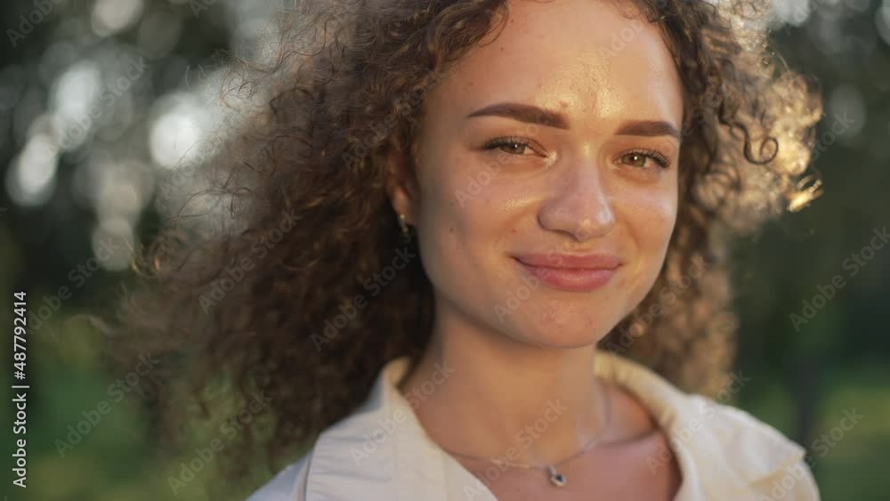 Headshot gorgeous Caucasian young woman posing in sunshine outdoors. Close-up portrait of confident beautiful brunette curly-haired lady looking at camera smiling in sunrays. Slow motion Headshot gorgeous Caucasian young woman posing in sunshine outdoors. Close-up portrait of confident beautiful brunette curly-haired lady looking at camera smiling in sunrays. Slow motion