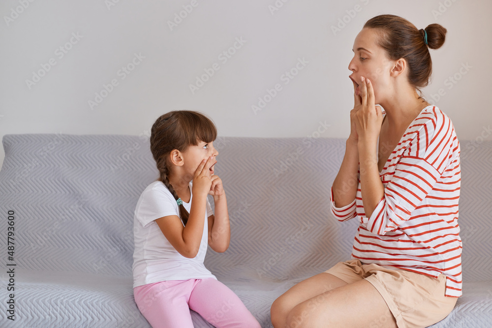 Portrait of young adult Caucasian woman wearing striped shirt and shorts sitting on sofa with little girl, female and kid doing exercise for restructuring oral muscular phonetic targets. Portrait of young adult Caucasian woman wearing striped shirt and shorts sitting on sofa with little girl, female and kid doing exercise for restructuring oral muscular phonetic targets.
