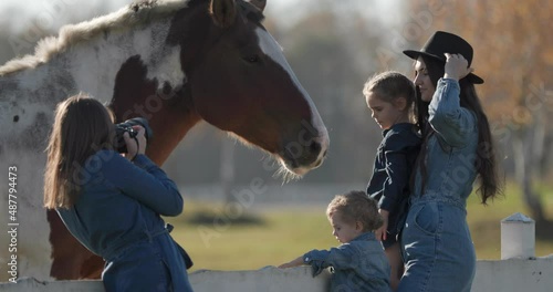 Female photographer is taking photos of family and a horse on a medow with a white wooden fence.  Family photo shoot with with kids. Sunny, bright day. 4k  60p to 23.976 slow motion. Telephoto shoot.