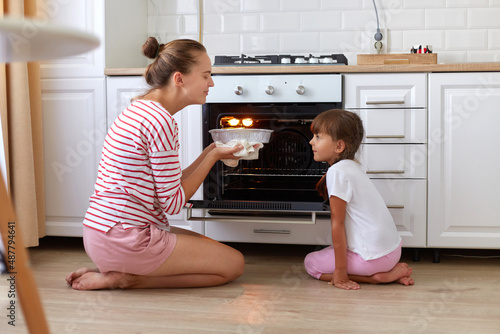 Happy young mum and cute kid daughter baking pie or cake in kitchen, mother and child smelling delicious tasty dessert, people sitting on floor, enjoying flavor.