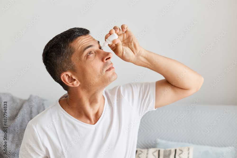 Indoor shot of brunette Caucasian man applying eyes drops, having problems with eyesight, treatment, overworked male using eyedropper for treating eyes. Indoor shot of brunette Caucasian man applying eyes drops, having problems with eyesight, treatment, overworked male using eyedropper for treating eyes.
