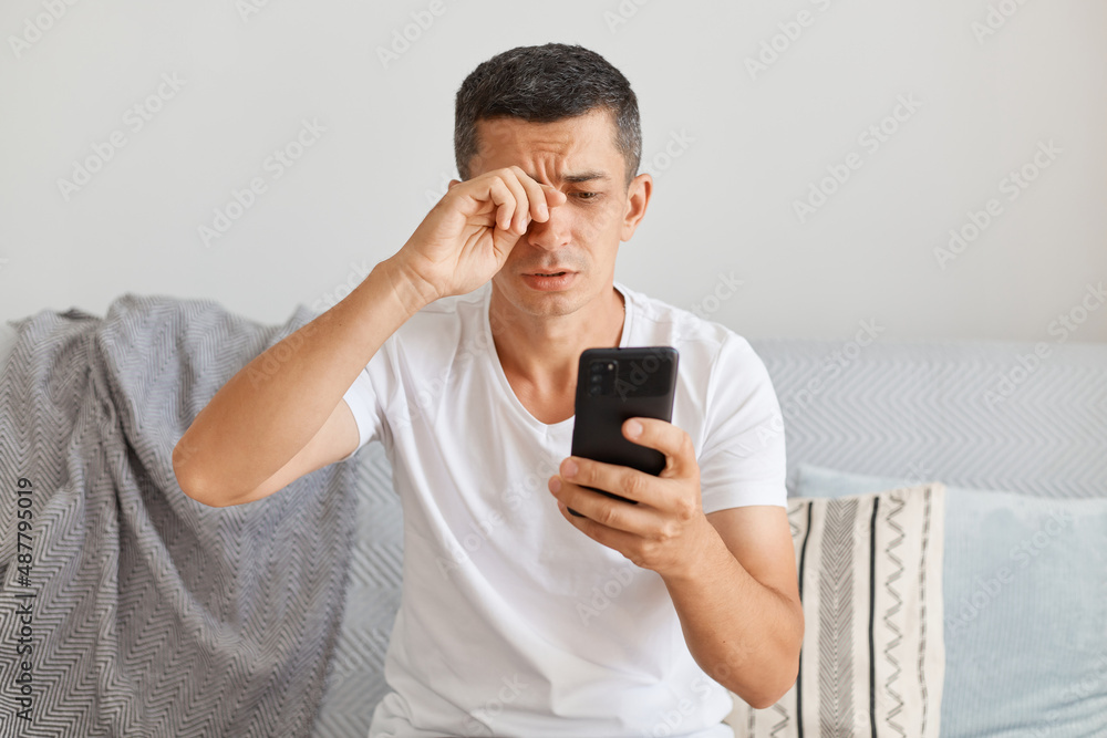 Portrait of tired upset man wearing casual style white t shirt sitting on cough, using smart phone, looks exhausted, rubbing eye, looking at gadget screen with sorrow. Portrait of tired upset man wearing casual style white t shirt sitting on cough, using smart phone, looks exhausted, rubbing eye, looking at gadget screen with sorrow.
