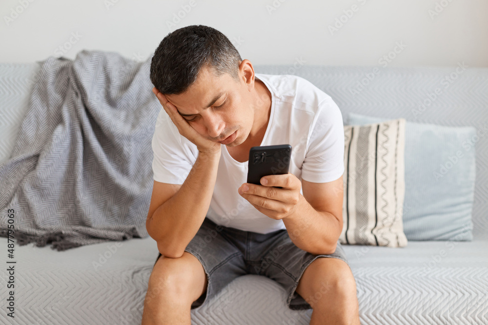 Portrait of exhausted sleepy bored man wearing casual style white t shirt sitting on cough, keeps eyes closed, having nap, holding cell phone in hands, looks tired.