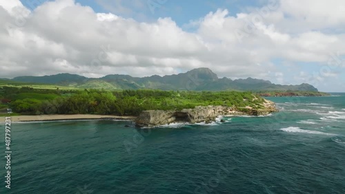 Slow wide drone aerial tracking shot of the tip of Shipwreck Beach, waves crashing against rocks. Large clouds in sky, Mountains in the background photo