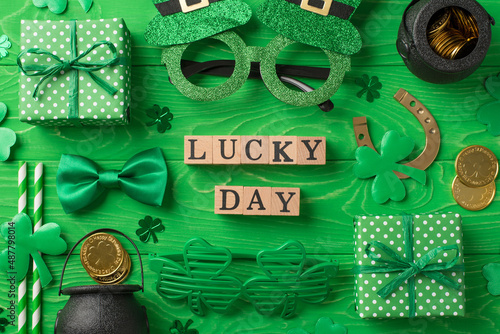 Top view photo of st patricks day decor wooden cubes labeled lucky day party glasses straws green bow-tie gift boxes horseshoe clovers pots with gold coins on isolated green wooden table background