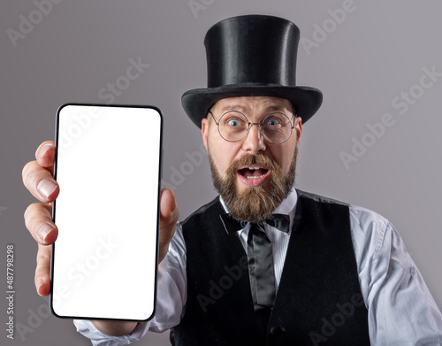 Classy bearded gentleman in suit and top hat standing over grey background and showing modern smartphone with white empty screen. Facial expressions, copy space.