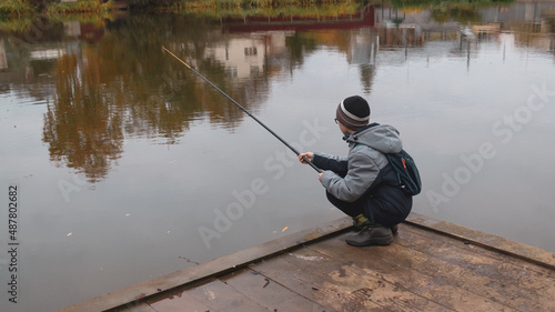 A teenage Caucasian boy is fishing on a calm autumn day on the lake squatting