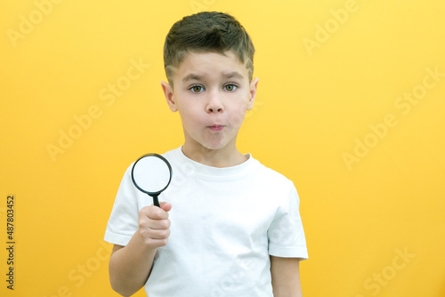 Positive curious schoolboy in casual clothes looks at the camera through a magnifying glass, on a yellow background.