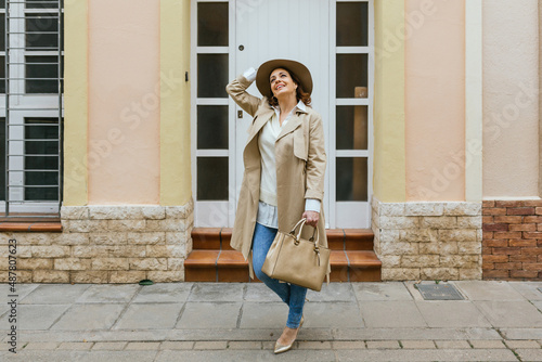 An adult woman in front of her front door posing with her hat and purse while looking up