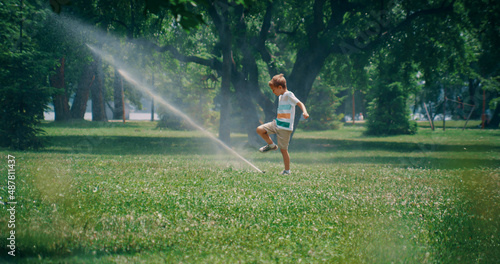 Playful elementary age boy kicking water sprinkler jet. Kid enjoy time in park
