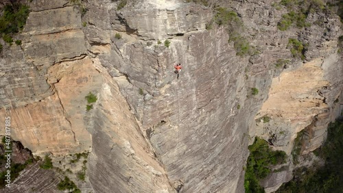 Rock climbers scaling an incredible huge steep mountain above a green valley. He slowly struggles and makes his way up the mountain face. Aerial drone shot.