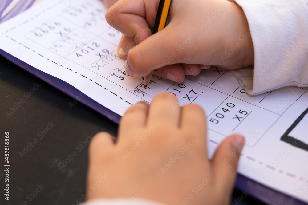 Close up on little girl hands doing school homework. Student writing with pencil maths multiplication exercise sheet - obrazy, fototapety, plakaty 